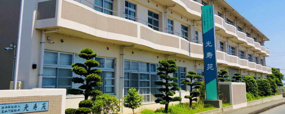 介護施設の建物外観。3階建てで、白と黄色系の外壁、大きな窓が特徴。植栽で整備された外構。